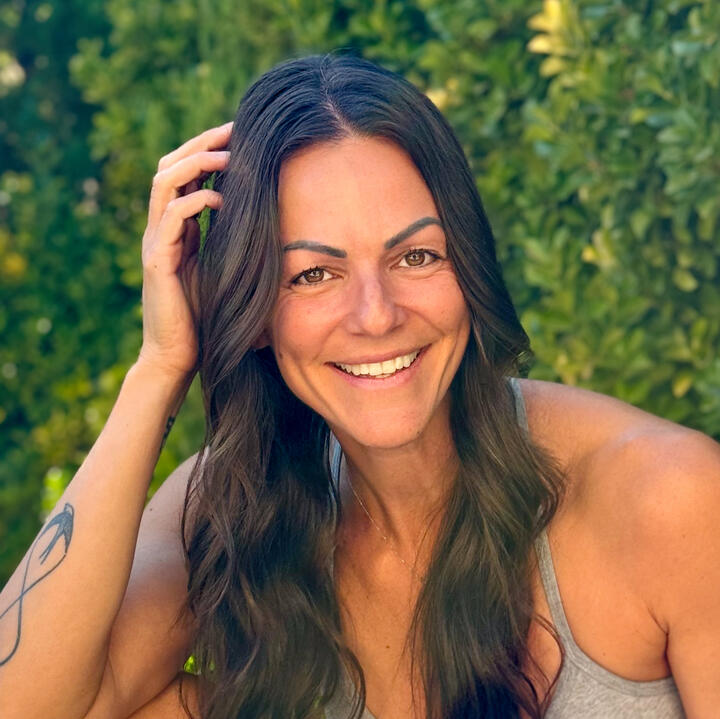 Danielle with brown eyes and brown straightened chest-length hair standing against a gray background, wearing a black v-neck tank top with crystal necklace